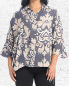 A woman wearing a loose, short-sleeve button-up blouse with a gray and beige damask pattern, paired with black pants, standing against a white brick wall.