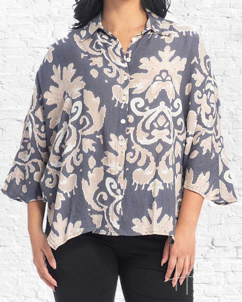 A woman wearing a loose, short-sleeve button-up blouse with a gray and beige damask pattern, paired with black pants, standing against a white brick wall.