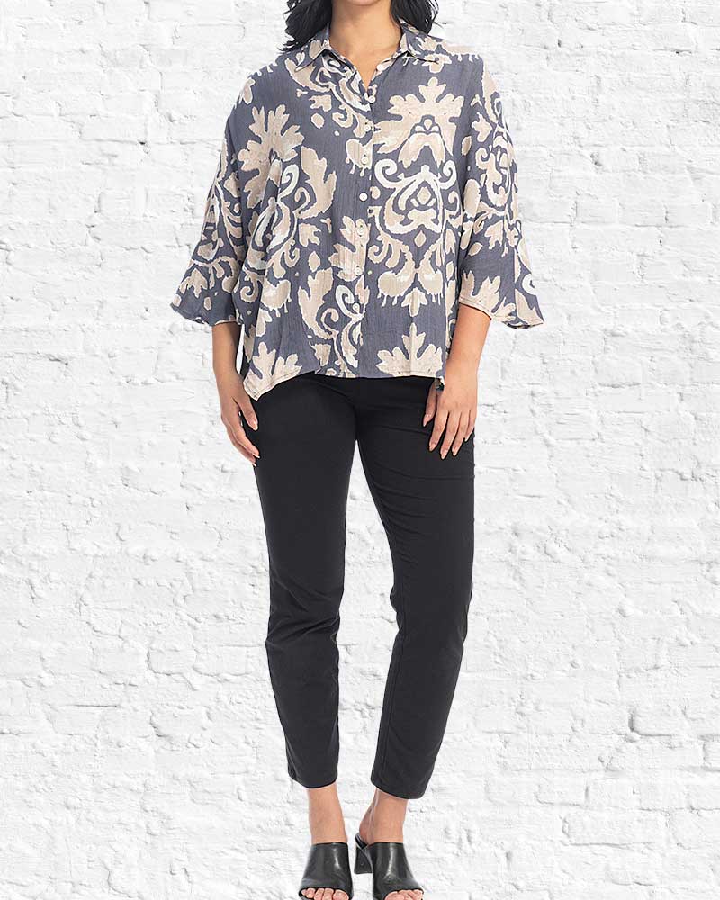 A woman modeling a loose, short-sleeve button-up blouse with a beige and slate gray damask pattern, paired with black cropped pants and black slide sandals, standing against a white brick wall.