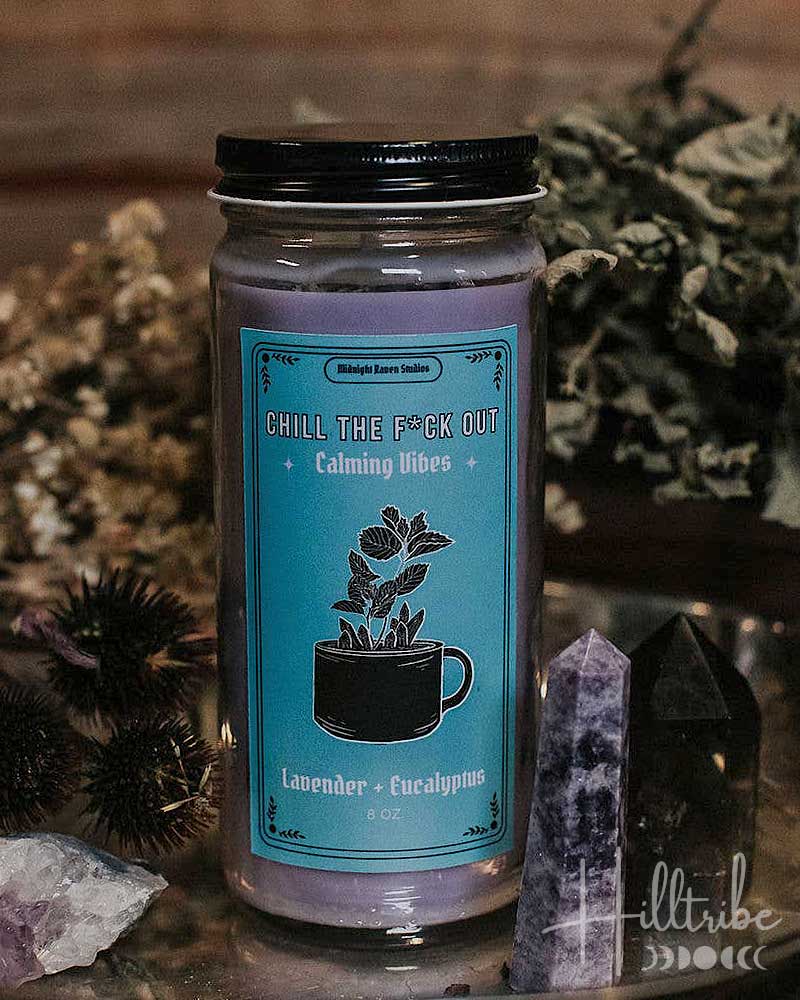 Candle jar with a blue label featuring text and graphics, surrounded by dried plants and crystals.