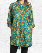 A woman wearing a teal green tunic top with a colorful floral and leaf pattern, paired with black pants, standing against a white brick wall.