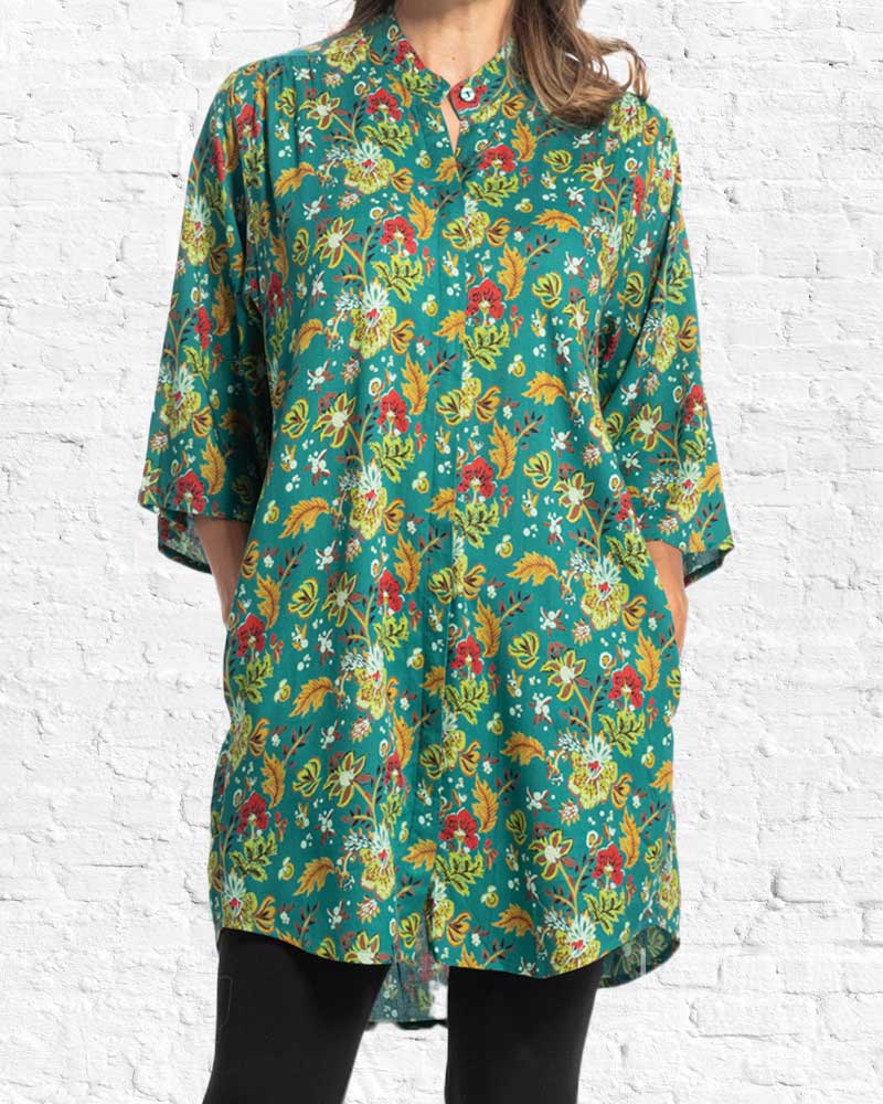 A woman wearing a teal green tunic top with a colorful floral and leaf pattern, paired with black pants, standing against a white brick wall.