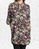 A woman wearing a black three-quarter sleeve tunic top with a dense floral and butterfly print in cream, purple, and brown, standing against a white brick wall.