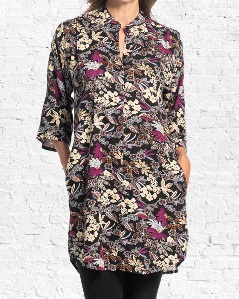 A woman wearing a black three-quarter sleeve tunic top with a dense floral and butterfly print in cream, purple, and brown, standing against a white brick wall.