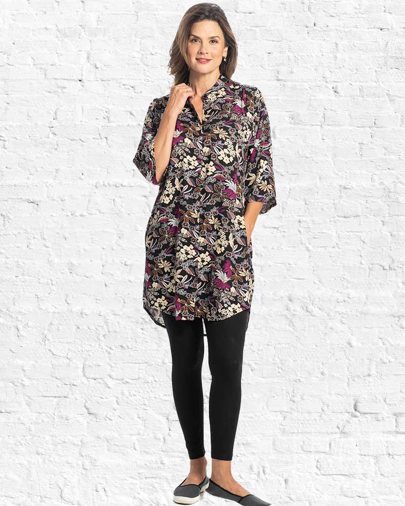 A woman wearing a knee-length black tunic dress with a dense multicolour floral and butterfly print in purple, beige, and brown, set against a white brick wall background.