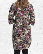 A woman viewed from the back wearing a knee-length black tunic dress with a dense multicoloured floral and butterfly print in purple, beige, and brown, set against a white brick wall background.