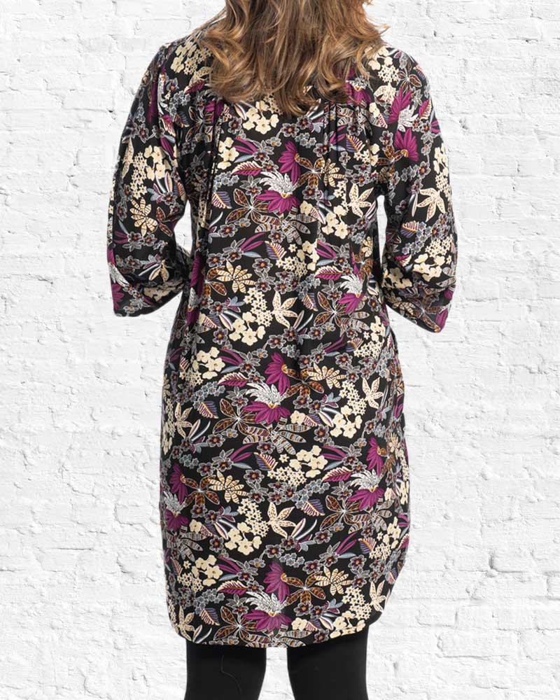 A woman viewed from the back wearing a knee-length black tunic dress with a dense multicoloured floral and butterfly print in purple, beige, and brown, set against a white brick wall background.