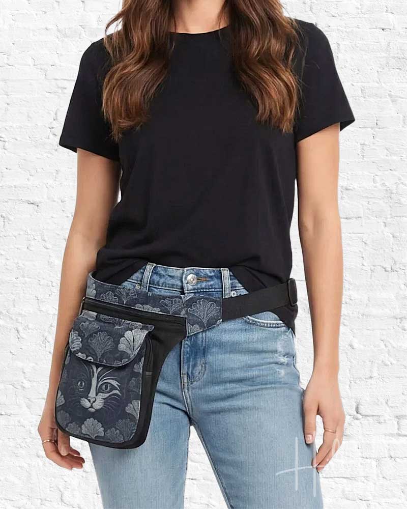 Person wearing a black t-shirt and blue jeans with a cat-themed waist bag against a white brick wall.