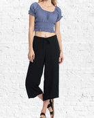 Woman wearing a blue top and black pants against a white brick wall.