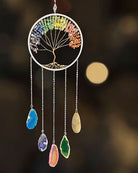 A circular chakra tree wind chime with colorful gemstone accents and five dangling teardrop-shaped crystals on silver chains, set against a dark blurred background with warm bokeh lights.