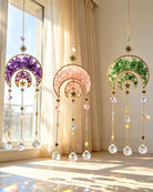 Three hanging decorative suncatcher ornaments with crescent moon and star designs, shown in purple, pink, and green crystal-filled halves, suspended in front of a sunlit window with curtains.