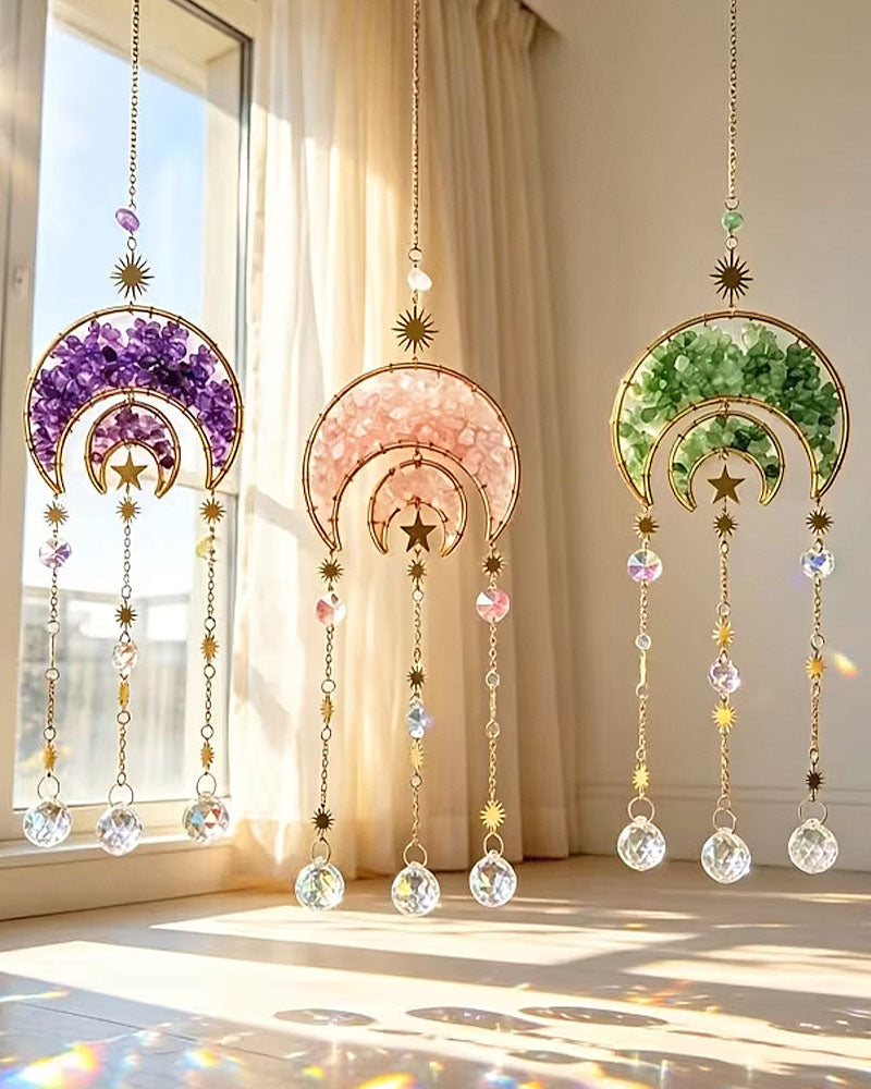 Three hanging decorative suncatcher ornaments with crescent moon and star designs, shown in purple, pink, and green crystal-filled halves, suspended in front of a sunlit window with curtains.