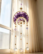 A gold crescent moon and star window hanging ornament with purple crystal accents and dangling clear crystal prisms, displayed in front of a window with beige curtains.
