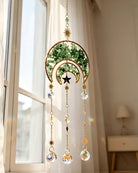 A gold crescent moon and star suncatcher with green leaves and hanging clear crystal prisms, hanging in front of a window in a softly lit room.