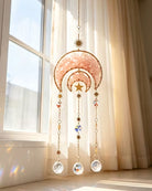 A gold-toned crescent moon and star suncatcher with pink crystal and faceted beads hangs in front of a sunlit window with sheer curtains.