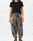 Person wearing a black t-shirt and colorful patchwork harem pants against a white brick wall.