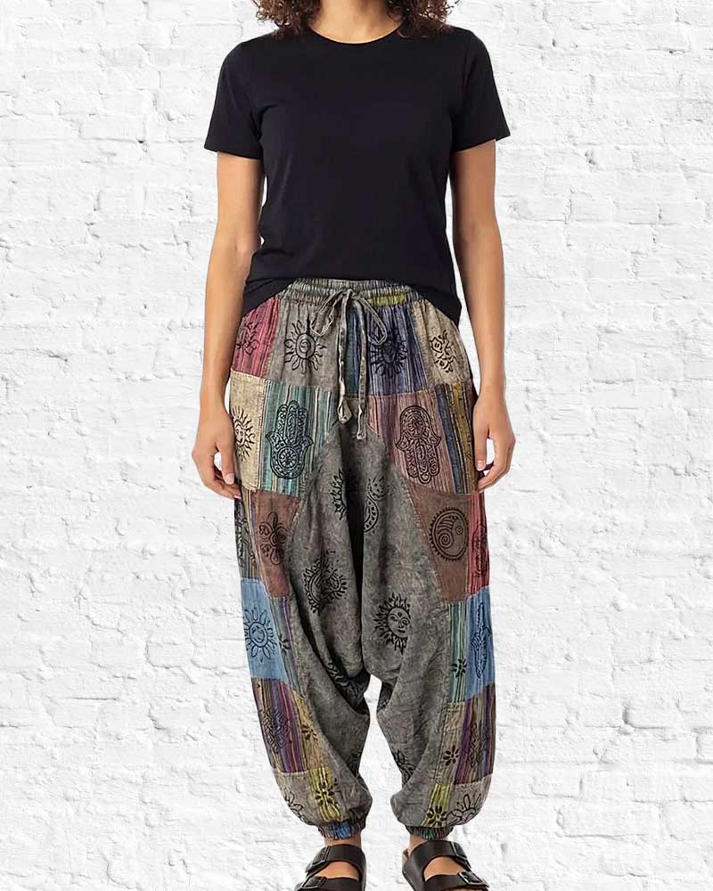 Person wearing a black t-shirt and colorful patchwork harem pants against a white brick wall.