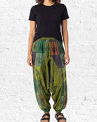 Person wearing a black t-shirt and green patterned harem pants against a white brick wall.