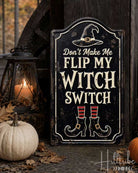 A decorative Halloween sign with the text 'Don't Make Me Flip My Witch Switch' in distressed cream lettering on a black background, featuring a witch hat and boots illustration. The sign is styled outdoors on a stone surface with autumn leaves, a small white pumpkin, an orange pumpkin, a lantern, a broom, and a black pot.