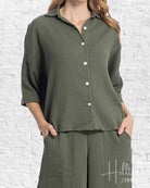 Person wearing a green button-up shirt and wide-leg pants against a white brick wall.