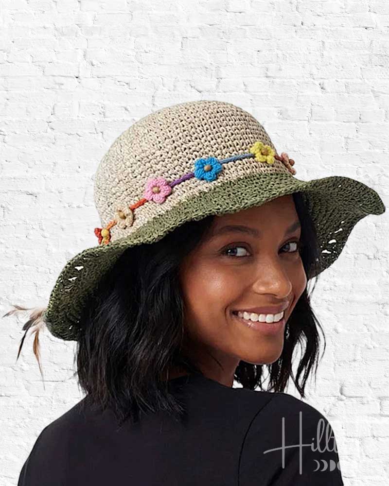 Woman wearing a crocheted hat with colorful flower decorations against a white brick wall.