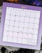 A vertical monthly calendar page for April, featuring a fantasy-style illustration of two cats around a campfire under a full moon on a purple background. The top section shows the illustrated scene, and the bottom section shows a grid layout for days of the week with blank spaces for writing and a small monthly calendar inset at the bottom right.