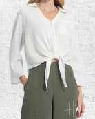 Person wearing a white blouse with a tie detail and green pants against a white brick wall.