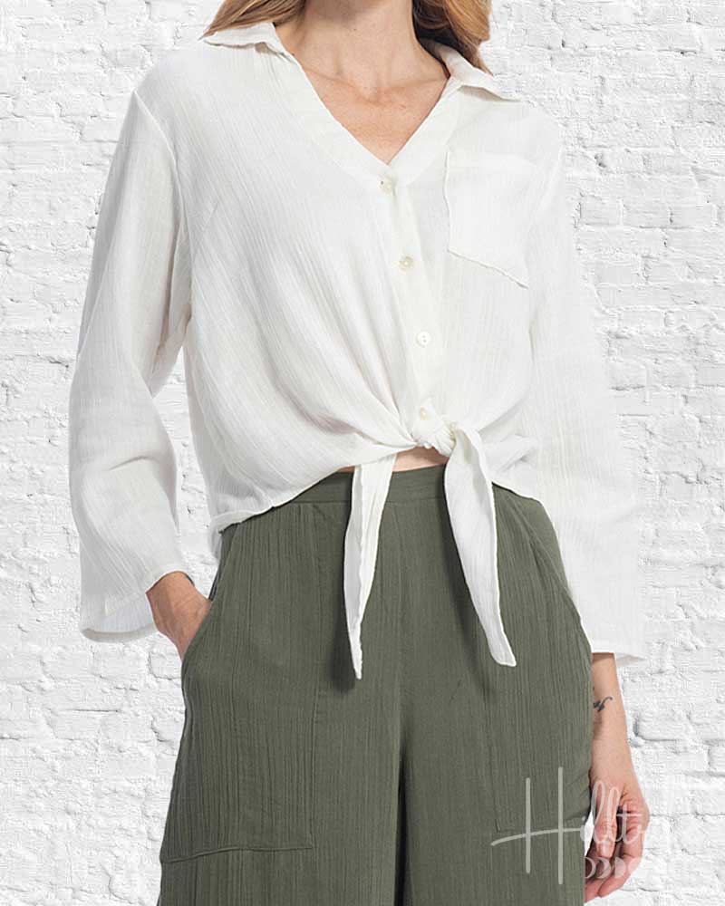 Person wearing a white blouse with a tie detail and green pants against a white brick wall.