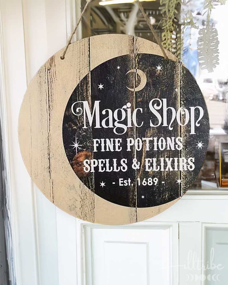 A round hanging wooden sign with a distressed wood-grain border and a dark central panel reading 'Magic Shop Fine Potions Spells & Elixirs Est. 1689' in white lettering, suspended by twine in a room setting.