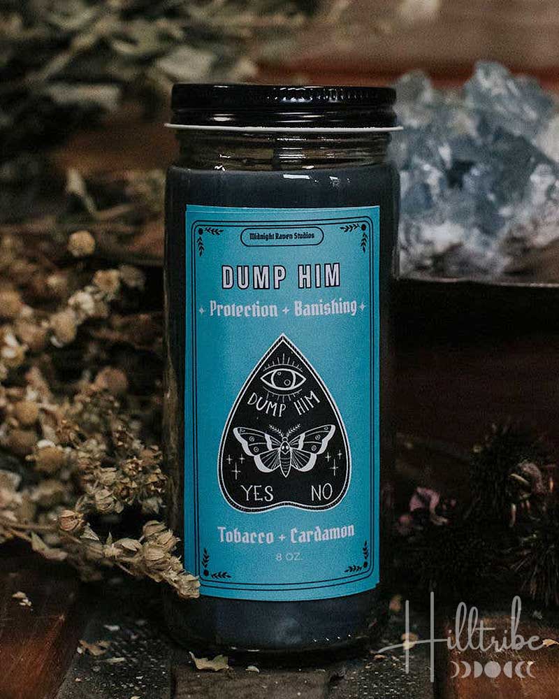 A glass jar candle with a black lid and a turquoise label reading 'DUMP HIM' and 'YES NO', styled on a rustic wooden surface with dried herbs and a crystal in the background.
