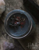 A small glass jar filled with dark gray wax and topped with dried red flowers, styled on a textured stone surface with crystals and a black crystal point.