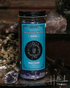 A jar candle labeled 'Intuitive Witch' and 'Berry Vanilla' sits on a rustic metal tray with crystals and dried botanicals against a dark wooden background.
