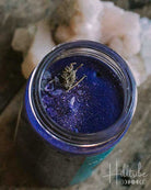 A small open jar filled with deep blue glittery wax or resin, placed on a distressed wooden surface next to a pale pink crystal, in a moody dark setting.