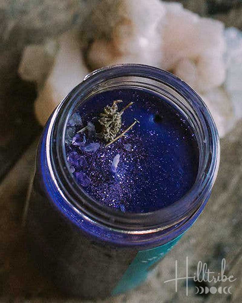 A small open jar filled with deep blue glittery wax or resin, placed on a distressed wooden surface next to a pale pink crystal, in a moody dark setting.