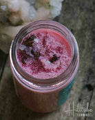 A small open jar filled with pink wax melts sits on a rustic wooden surface next to a pale crystal cluster. The scene is dimly lit with a moody, atmospheric background.