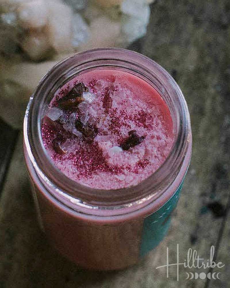 A small open jar filled with pink wax melts sits on a rustic wooden surface next to a pale crystal cluster. The scene is dimly lit with a moody, atmospheric background.