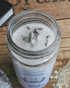 A glass jar candle with white wax and dried botanicals on top, styled on a rustic wooden surface with crystals, dried flowers, and a book.