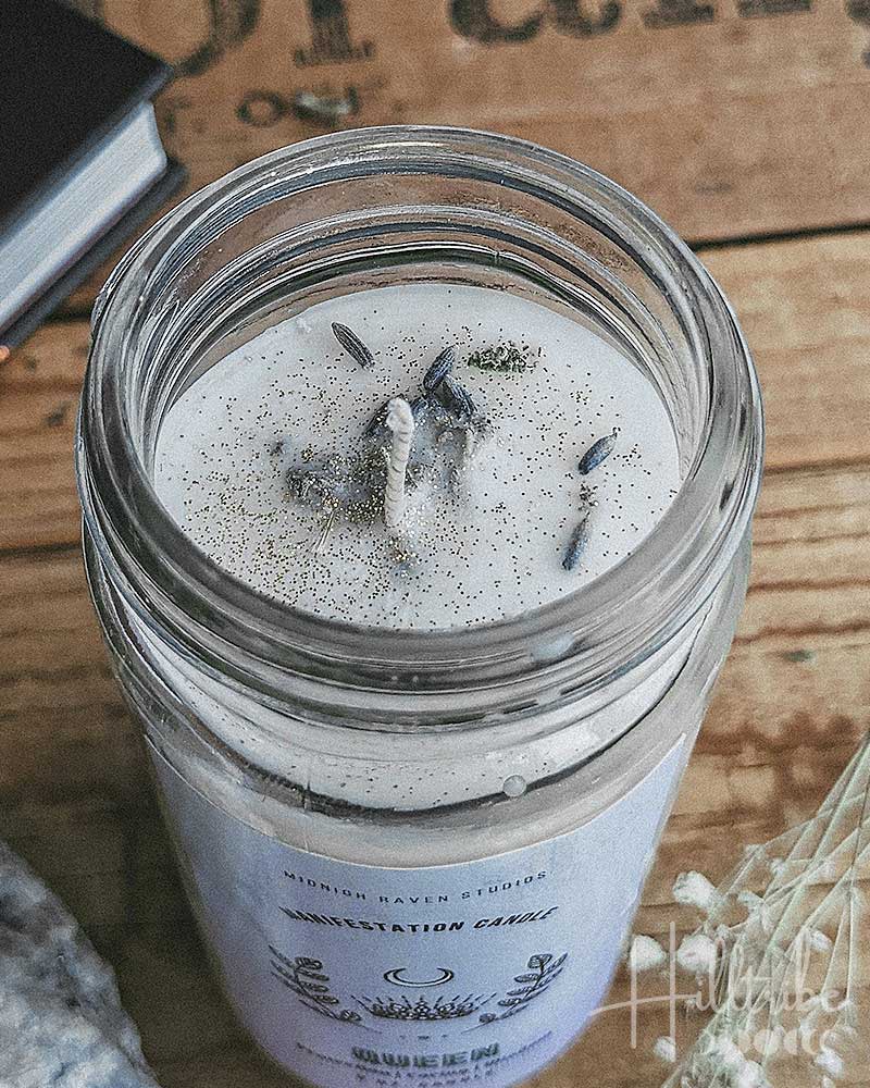 A glass jar candle with white wax and dried botanicals on top, styled on a rustic wooden surface with crystals, dried flowers, and a book.