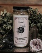 A glass jar candle labelled 'Rebel' styled on a dark tray with dried herbs, crystals, and a rustic wooden background.