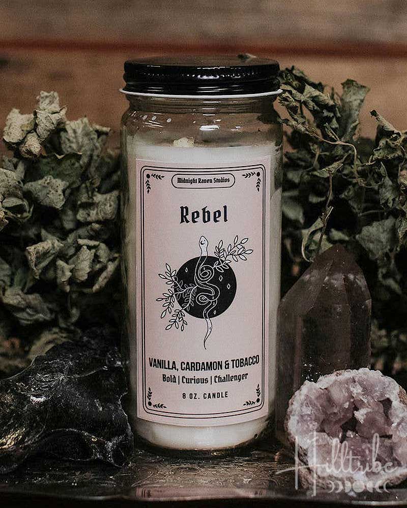 A glass jar candle labelled 'Rebel' styled on a dark tray with dried herbs, crystals, and a rustic wooden background.
