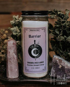 A jar candle labelled 'Warrior', placed on a dark tray with crystals and dried botanicals against a rustic wooden background.