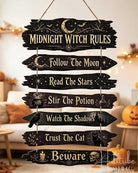 A rustic hanging wooden sign with seven arrow-shaped planks reading 'Midnight Witch Rules' and related phrases, shown in a cozy Halloween-themed living room with pumpkins and string lights.