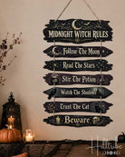 A rustic multi-tiered wooden sign with the text 'Midnight Witch Rules' and seven spooky phrases, hanging on a beige wall above a decorated mantel with pumpkins, candles, and black lace.
