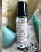 A 10ml roll-on bottle of 'Money Magic' crystal infused ritual oil by The Patchouli Aventuring Arts, styled with a black cap, white label, and surrounded by green leaves, a turquoise stone, and a green candle on a white faux fur background.