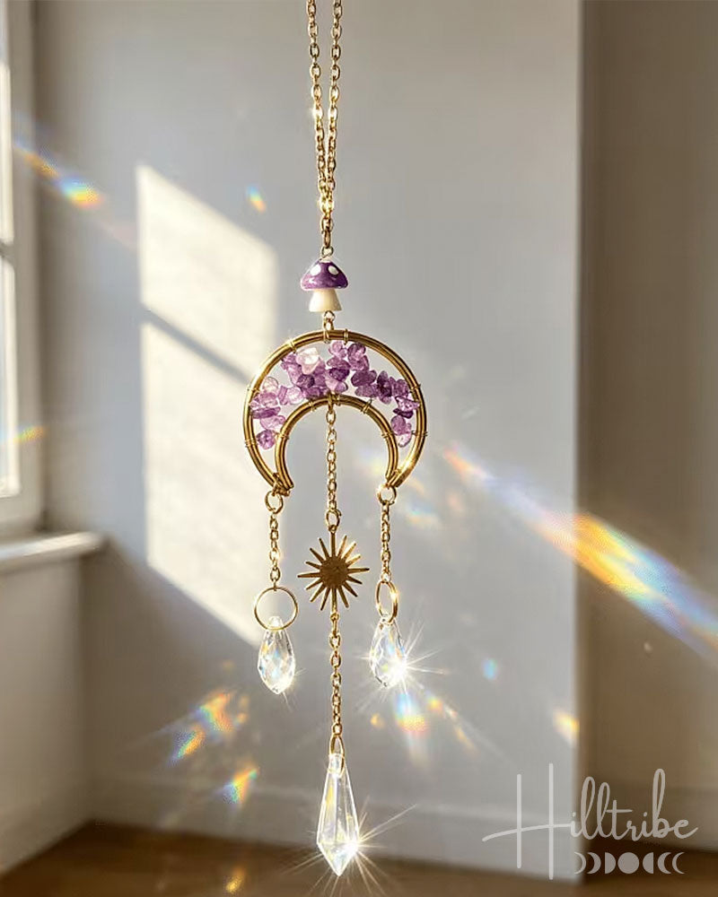 A gold crescent moon and sun pendant ornament with purple crystals and clear hanging prisms, suspended in a sunlit room with rainbow light leaks.