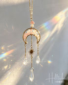 A gold-toned crescent moon and star pendant necklace with pink crystal inlay and dangling clear teardrop crystals, photographed against a sunlit neutral background with rainbow light leaks. The Hilltribe logo appears in the lower right corner.