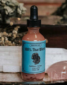 A small amber dropper bottle labelled '100% That Whiskey' with a turquoise label and black dropper cap, placed on a tray with dried herbs, white crystals, and a pink stone against a rustic wooden background.