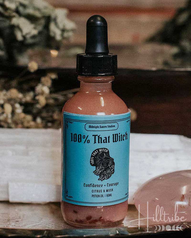 A small amber dropper bottle labelled '100% That Whiskey' with a turquoise label and black dropper cap, placed on a tray with dried herbs, white crystals, and a pink stone against a rustic wooden background.