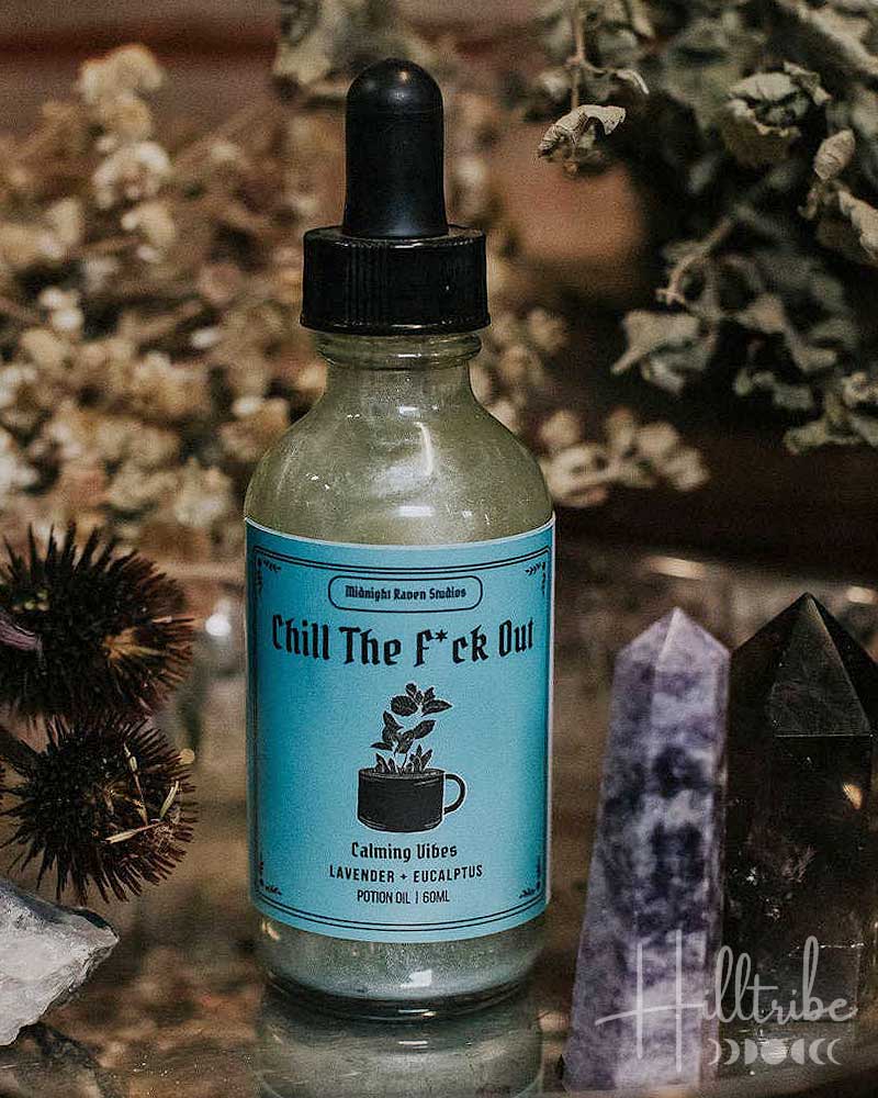 A small glass dropper bottle labelled 'Chill The F*ck Out' sits on a reflective metal tray surrounded by dried flowers, crystals, and a rustic wooden background.