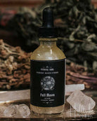 A small glass dropper bottle labelled 'Full Moon' sits on a rustic wooden surface with crystals, dried herbs, and a blurred wood background.